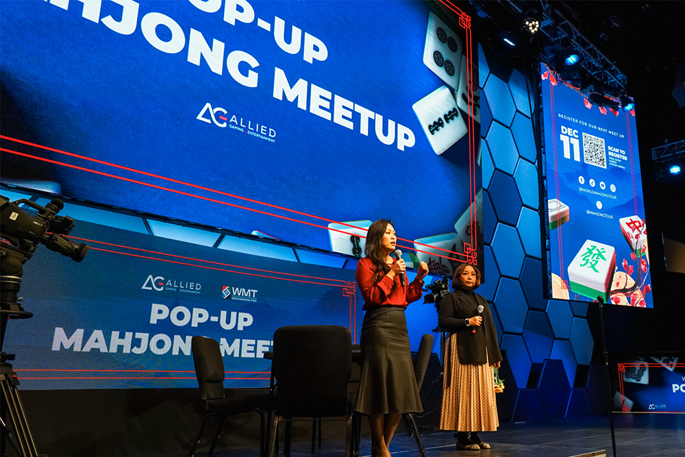 Speakers on stage at a pop-up Mahjong meetup, large blue screen behind them.