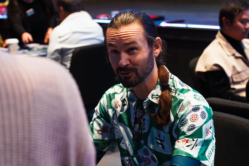 Bearded man with long hair in a tropical shirt sits at a conference table, looking ahead.