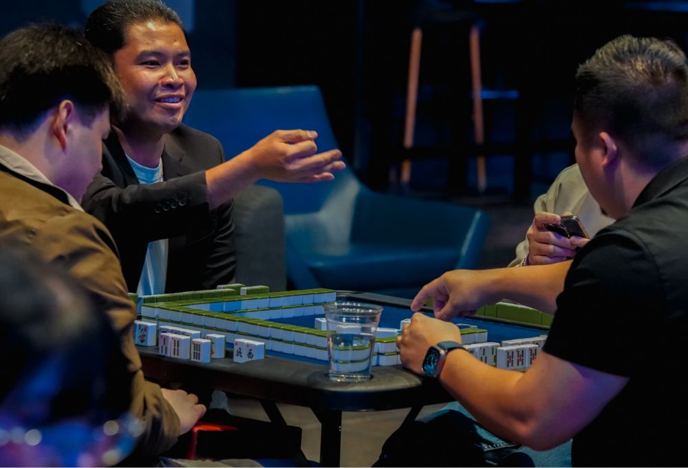 A group of players around a mahjong table, one man smiling and gesturing.
