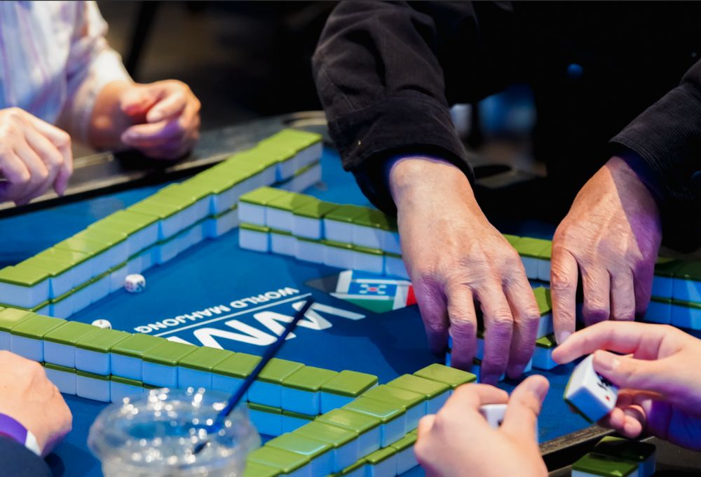 Several hands reach for tiles around a mahjong table on a blue surface.