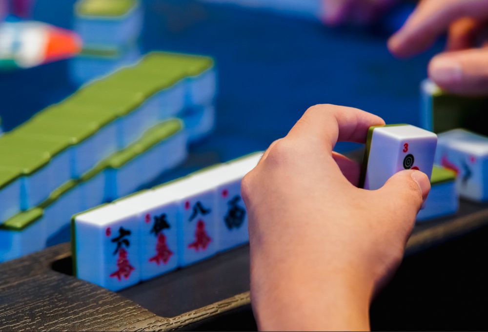 A hand places a Mahjong tile over a row of tiles on the blue gaming table.