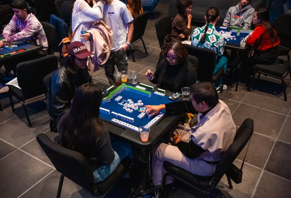 Several players around a blue mahjong table; tiles arranged in play, focused faces.