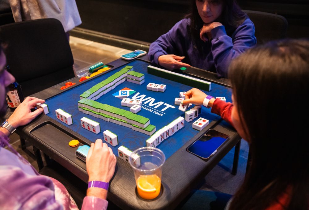 Mahjong in progress at a teal-green table; players' hands and tiles laid out, orange juice glass nearby.